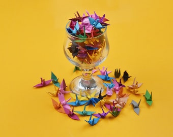 May include: A glass vase filled with colorful origami paper cranes. The cranes are in various shades of pink, blue, green, yellow, and orange. Some cranes are scattered around the vase on a yellow surface.