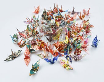 May include: A collection of colorful origami paper cranes, folded in a traditional Japanese style, are arranged in a circle around a glass jar. The cranes are made from patterned paper with various colors and designs.