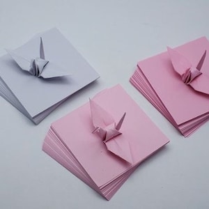 May include: Three stacks of origami paper, each with a folded origami crane on top. The stacks are white, pink, and pink.