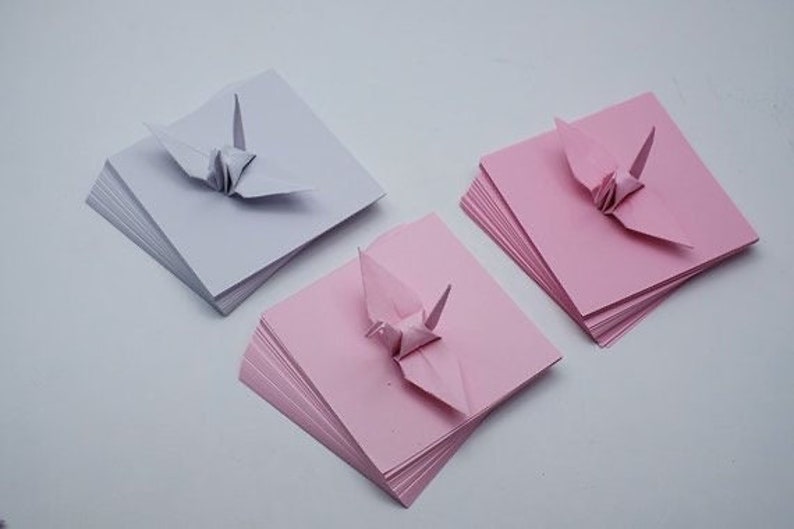 May include: Three stacks of origami paper, each with a folded origami crane on top. The stacks are white, pink, and pink.
