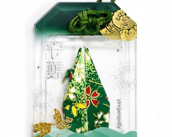 May include: A rectangular clear acrylic omamori charm featuring a green fabric insert with a floral pattern. The charm includes a green cord, a gold metal accent, and the text PROTECTION and yingporipolly printed on the front surface.