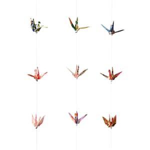 Könnte beinhalten: Neun bunte Origami-Kraniche hängen an Fäden vor einem weißen Hintergrund. Die Kraniche sind aus gemustertem Papier gefertigt und in verschiedenen Höhen aufgehängt.