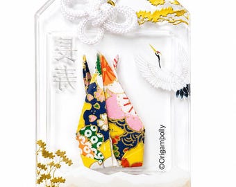May include: A clear rectangular omamori charm featuring a colorful patterned paper crane at the center. The charm includes a white cord, a white crane illustration, gold leaf accents, Japanese characters, the text "HEALTH & LONGEVITY", and "©Origampolly".