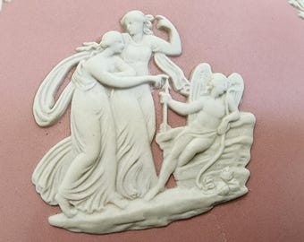 Puede incluir: Un plato decorativo ovalado rosa con un diseño en bajorrelieve blanco. El relieve representa tres figuras y un ángel. El plato está bordeado con un diseño floral blanco. Las figuras visten atuendo clásico.