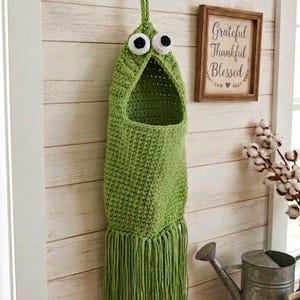 May include: A green crocheted plastic bag holder featuring a wide, open mouth and two large, round white and black eyes at the top. The cylindrical body is made of textured yarn and finishes with a long, straight fringe at the bottom edge.