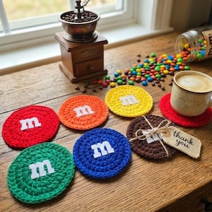 May include: A set of six round, crocheted coasters in red, orange, yellow, green, blue, and brown. Each coaster features a white felt letter m in the center. One brown coaster is tied with twine to a small beige tag with the text thank you.