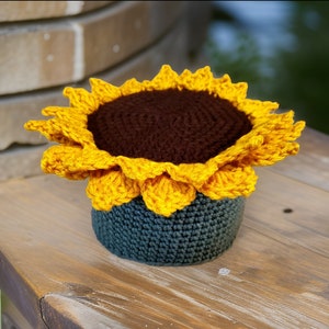 May include: A crocheted sunflower-shaped planter with a dark brown center and yellow petals. The planter is made of a dark blue yarn.