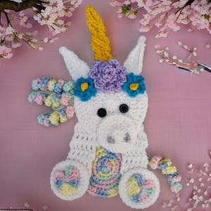 May include: A handmade crochet unicorn with a yellow horn, white body, and colorful mane and tail. The unicorn has a purple flower crown with blue and yellow accents. Black button eyes and a gray nose complete the design. The unicorn is sitting on a pink surface.