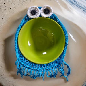 May include: A handmade blue crochet bowl cozy with white and black eyes, holding a green bowl. The cozy has a fringed edge, resembling a cartoon character. The bowl is centered within the cozy.