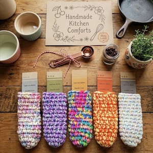 May include: A collection of colorful, handmade crochet cast iron skillet handle covers. The covers are in various colors, including pink, purple, orange, and white. The image also includes a sign that says "Handmade Kitchen Comforts".