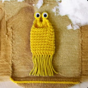 May include: A yellow crocheted monster with googly eyes and a fringe. The monster is sitting on a brown knitted surface.