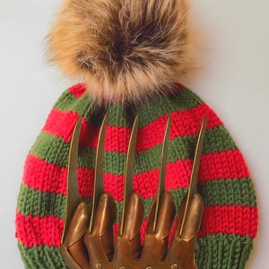 May include: A striped red and green knit beanie with a brown faux fur pom-pom. A brown leather glove with metal claws is in front of the beanie. The glove has five long, curved metal blades.