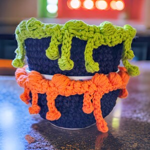 May include: Two stacked, dark blue ceramic bowls with crocheted green and orange accents. The green crochet resembles dripping slime around the top rim, while the orange crochet is around the bottom rim. The bowls are likely for Halloween or themed decor.
