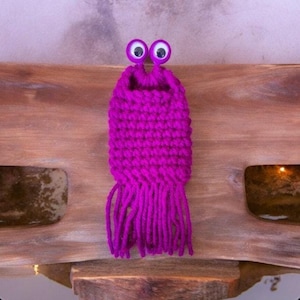May include: A small, crocheted purple monster with googly eyes and a fringe. The monster is made with yarn and has a pocket on the front.
