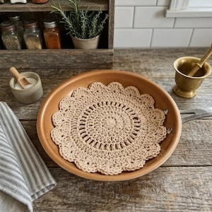 May include: A beige crochet doily sits inside a copper-colored frying pan on a wooden surface. A small ceramic bowl with a wooden spoon, a brass mortar and pestle, and a striped towel are also visible. A wooden shelf with jars of spices and herbs is in the background.