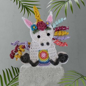 May include: A crocheted unicorn toy with a white body, rainbow mane and tail, and colorful flower accents. The unicorn is sitting on a gray stone surface.