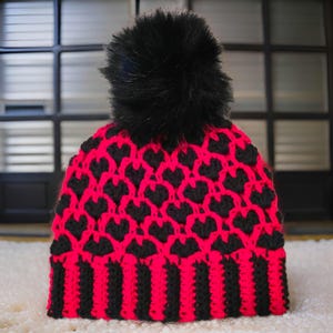 May include: A knitted winter hat with a black faux fur pom-pom. The hat features a pattern of black hearts on a bright pink background. The brim has alternating vertical stripes of black and pink.