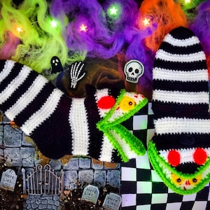 May include: A Halloween-themed crochet set featuring black and white striped leg warmers and a green and white monster-like design. The scene includes decorative elements like tombstones, a gate, and a sign that reads "No. 34".