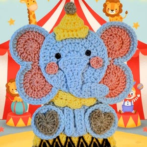 May include: A handmade crochet elephant toy in blue, pink, yellow, and brown. The elephant is wearing a yellow hat and collar, with a circus-themed background featuring a tent, stars, and cartoon animals. The elephant has large ears and a trunk.