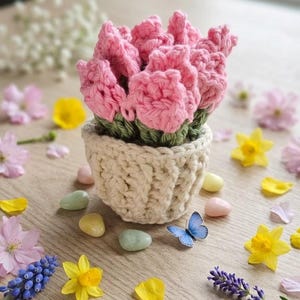 May include: A crocheted pink flower arrangement in a cream-colored pot. The flowers are surrounded by green leaves. The arrangement is on a wooden surface with scattered flowers, petals, and small decorative stones. A blue butterfly is also present.