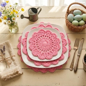 May include: A stack of three white square plates with pink crocheted doilies. A small basket of blue and green eggs sits to the right. A small watering can and a vase of flowers are also present.
