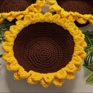 May include: Three round crochet bowls featuring a dark brown center and bright yellow petal-shaped edges that resemble sunflowers. The bowls are constructed with a dense, textured yarn stitch pattern, creating a deep, bowl-like shape for storage.