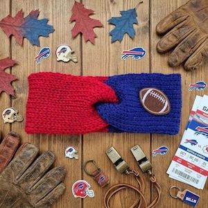 May include: A red and blue knit headband with a twisted center and a brown football patch, surrounded by brown leather gloves, autumn leaves, Buffalo Bills team pins, metal whistles, and a game day ticket for a Buffalo Bills versus Chiefs game.