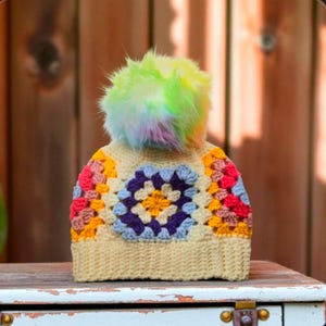 May include: A handmade crochet beanie with a colorful granny square design. The hat features a cream-colored base with a central square of blue, purple, and yellow. The edges are adorned with red, pink, and orange squares. A fluffy, multi-colored pom-pom tops the hat.
