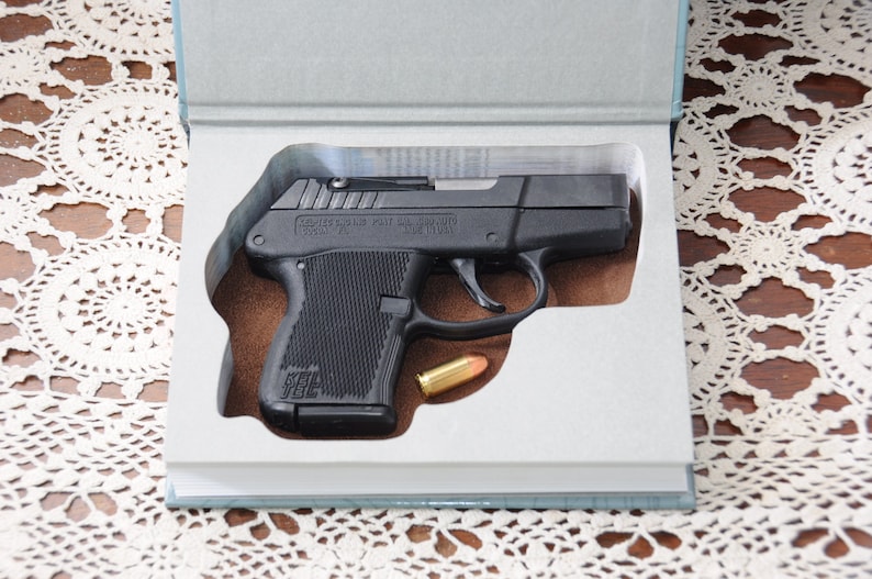 Op de afbeelding: Een zwart pistool met een zilveren slede, gepresenteerd in een boekvormige koffer. Er ligt een enkele kogel naast het pistool. De koffer is open en toont het vuurwapen en de kogel tegen een bruine achtergrond.