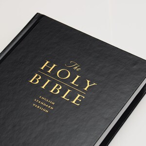 May include: A black hardcover book titled "The Holy Bible" with gold lettering. The text "English Standard Version" is also visible. The book is set against a white background.