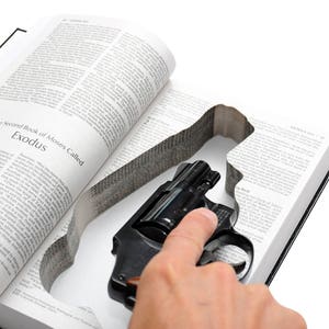 May include: A black revolver gun is nestled inside a hollowed-out book. The book is open to a page with the title "Exodus". A hand is touching the gun, suggesting it is being handled.