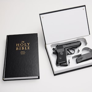 May include: A black handgun and magazine are displayed in a white-lined box next to a black book. The book is titled "The Holy Bible" in gold lettering. The box is open, revealing the gun.