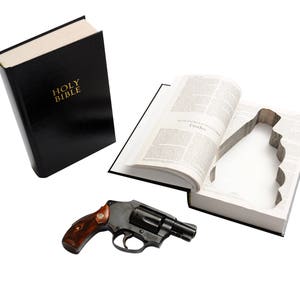 May include: A black Bible with gold lettering that reads "HOLY BIBLE" is next to an open book with a cutout. A black revolver with a wooden handle rests nearby. The items are on a white background.