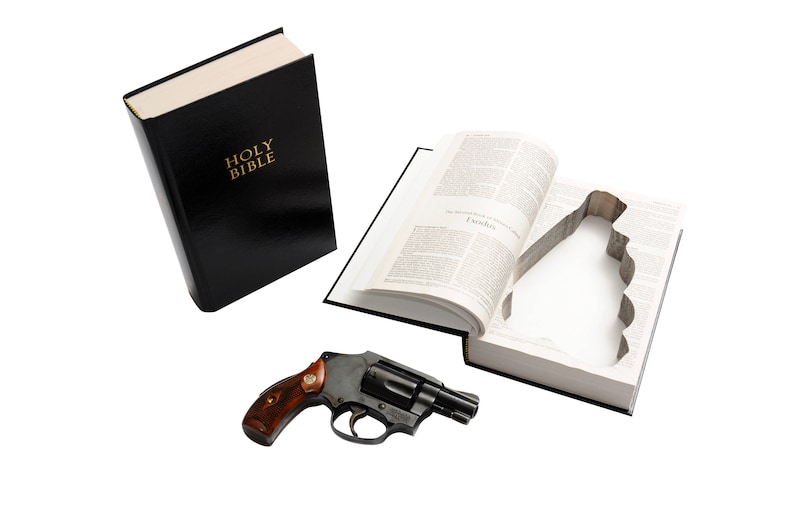 May include: A black Bible with gold lettering that reads "HOLY BIBLE" is next to an open book with a cutout. A black revolver with a wooden handle rests nearby. The items are on a white background.