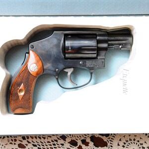 Op de afbeelding: Een zwarte revolver met een houten handvat, gepresenteerd in een witte doos. Het pistool heeft een zilveren trekker en een cilinder. Op de doos staat het woord "Etiquette".