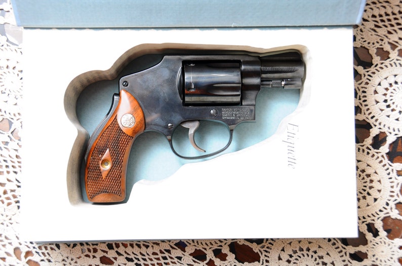 Op de afbeelding: Een zwarte revolver met een houten handvat, gepresenteerd in een witte doos. Het pistool heeft een zilveren trekker en een cilinder. Op de doos staat het woord "Etiquette".