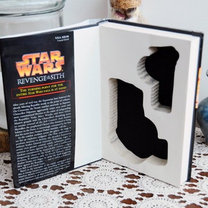 Op de afbeelding: Een Star Wars: Revenge of the Sith boek met een uitgesneden ontwerp. De kaft van het boek toont het Star Wars-logo en de titel. De uitsparing onthult een zwart interieur, waardoor een uniek display-item voor fans ontstaat.