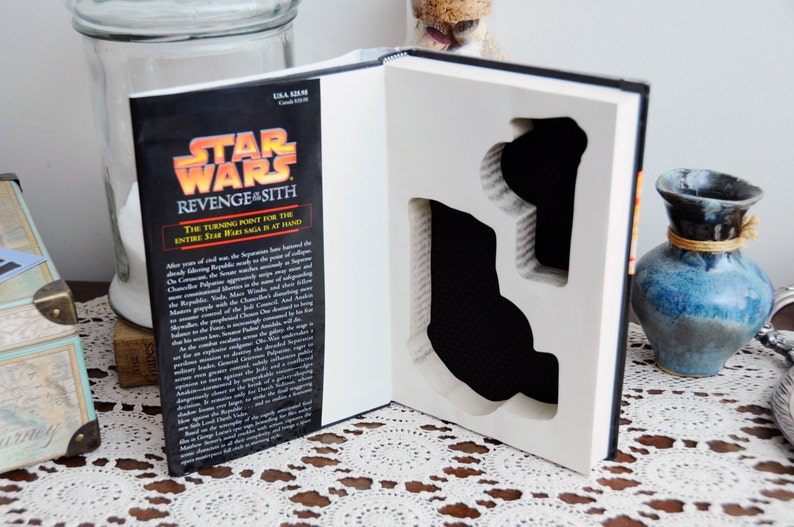 Op de afbeelding: Een Star Wars: Revenge of the Sith boek met een uitgesneden ontwerp. De kaft van het boek toont het Star Wars-logo en de titel. De uitsparing onthult een zwart interieur, waardoor een uniek display-item voor fans ontstaat.