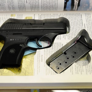 Op de afbeelding: Een zwart Ruger-pistool en magazijn liggen in een uitgesneden ruimte in een open boek. Het magazijn is metaalachtig met genummerde gaten. De pagina's van het boek tonen tekst en een kaart.