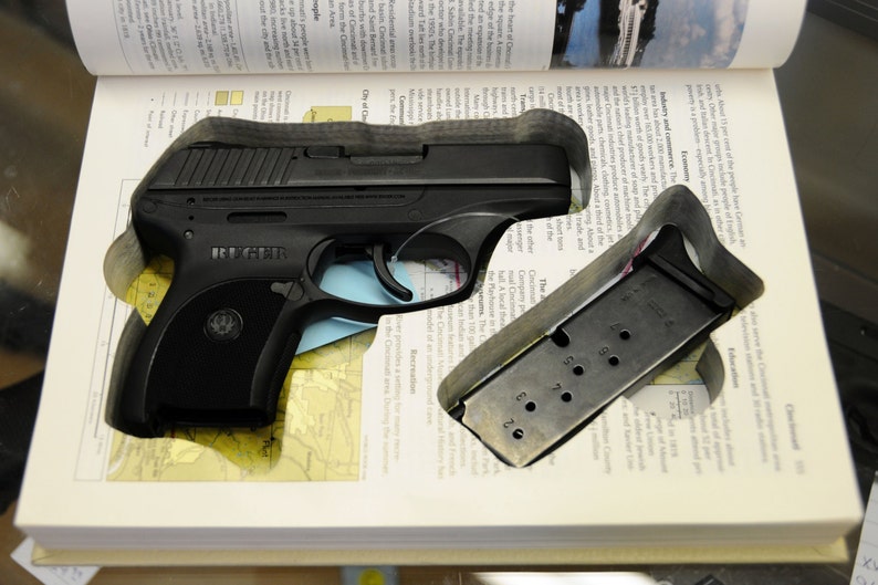 Op de afbeelding: Een zwart Ruger-pistool en magazijn liggen in een uitgesneden ruimte in een open boek. Het magazijn is metaalachtig met genummerde gaten. De pagina's van het boek tonen tekst en een kaart.