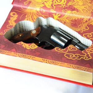Op de afbeelding: Een zwarte revolver ligt in een uitgehold boek. De kaft van het boek is rood met gouden draken- en wolkenpatronen. De revolver heeft een houten handvat en een zilveren loop.