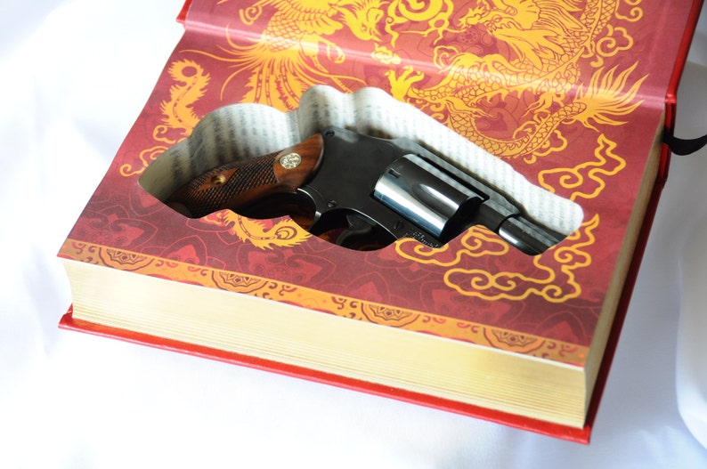 Op de afbeelding: Een zwarte revolver ligt in een uitgehold boek. De kaft van het boek is rood met gouden draken- en wolkenpatronen. De revolver heeft een houten handvat en een zilveren loop.