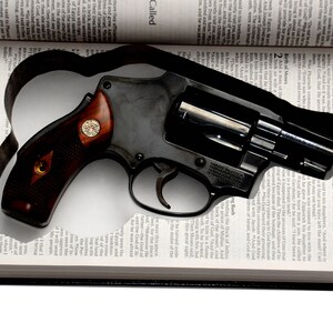 May include: A black revolver with a wooden handle rests on an open book. The book's pages are filled with text, and the revolver is positioned in the center. The handle has a gold emblem.