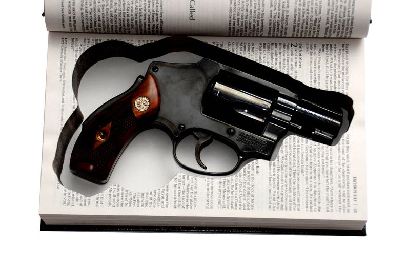 May include: A black revolver with a wooden handle rests on an open book. The book's pages are filled with text, and the revolver is positioned in the center. The handle has a gold emblem.