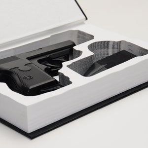 May include: A black handgun and magazine are nestled in a custom-cut compartment within a book. The book's pages are white, and the cover is black. The handgun is a dark, matte black.