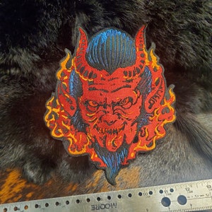 May include: A red and blue embroidered patch of a devil with flames around the head. The devil has a fierce expression and a pointed beard.