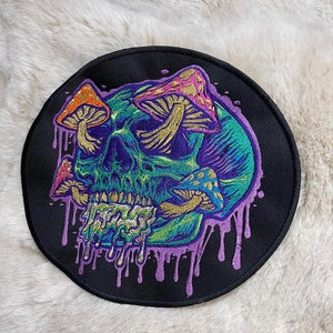 May include: A round, black patch with an embroidered design of a skull with mushrooms growing out of its eye sockets. The skull is dripping with purple and green liquid. The mushrooms are various colors, including orange, pink, and gold.
