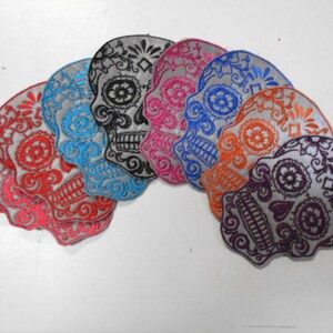 May include: A set of seven embroidered iron-on patches featuring colorful sugar skulls with intricate floral designs. The skulls are in various colors, including black, pink, blue, orange, and purple.