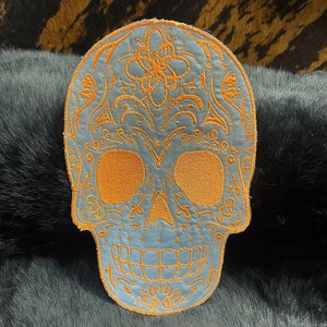 May include: A blue embroidered sugar skull patch with orange thread. The skull has intricate details and a floral design on the top of the head.