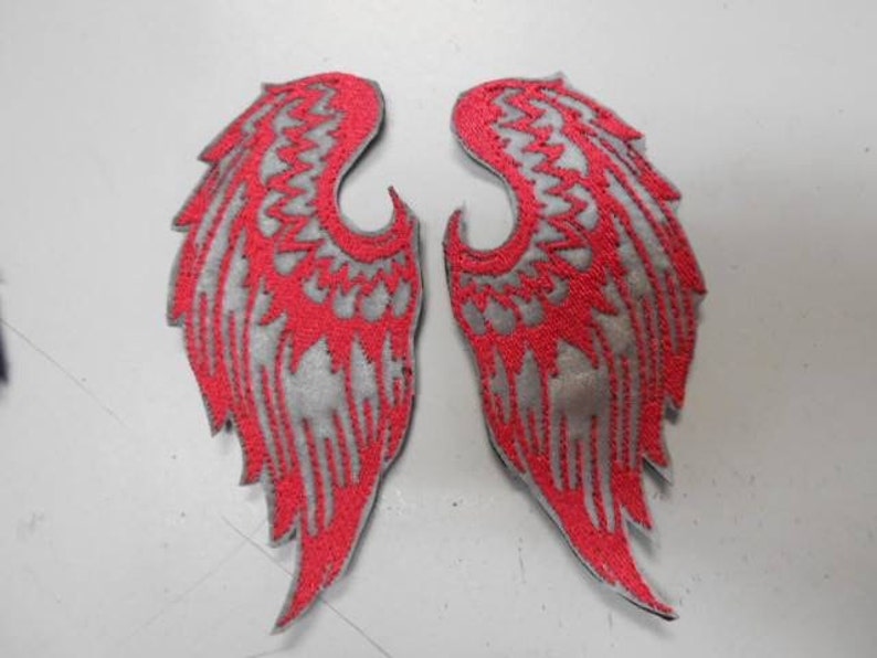 May include: Two red and gray embroidered angel wings patches. The wings are detailed with a zig-zag pattern and have a gray background with red stitching.